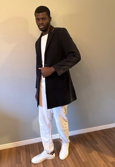 Abrigo negro con cuello marrón, camiseta blanca, pantalones de lino blancos y zapatillas blancas. El abrigo tiene un solo botón y un corte relajado.