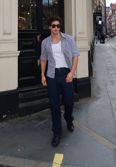 Un hombre viste una camisa de cuadros de manga corta y una camiseta blanca, con jeans oscuros y zapatos negros, camina por una calle de la ciudad.