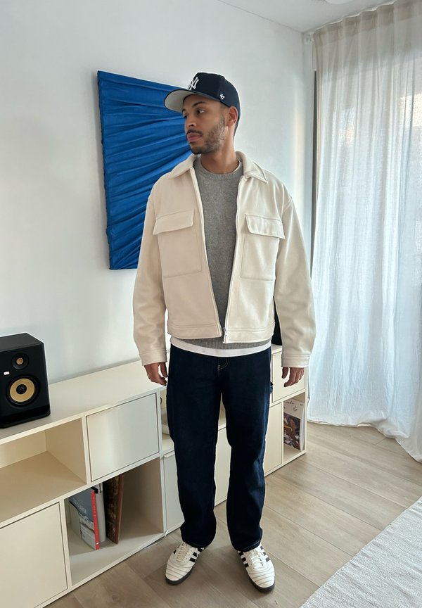 Giacca in pile bianca con due tasche frontali, maglione grigio, jeans blu e sneakers bianche con strisce nere. Sullo sfondo si trova un'opera d'arte su fondo blu.