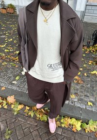 Casaco castanho, suéter creme com logotipo, calças castanhas e sapatos rosa claro. A cena inclui pavimento de paralelepípedos e folhas de outono.