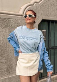 T-shirt di cotone azzurro con testo stampato, abbinato a una giacca di jeans blu e una mini gonna bianca. Gli accessori includono occhiali da sole quadrati bianchi.