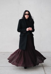 Manteau en laine noire porté sur un haut en maille noir, associé à une longue jupe maxi bordeaux fluide en tissu transparent superposé. Des lunettes de soleil noires complètent le look.