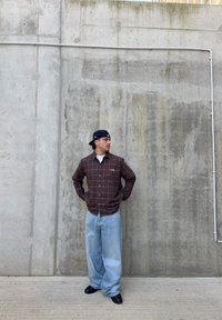 Braunes kariertes Hemd mit Knöpfen, hellblaue lockere Jeans und schwarze Schuhe. Betonwand im Hintergrund mit sichtbarer Textur und Linien.