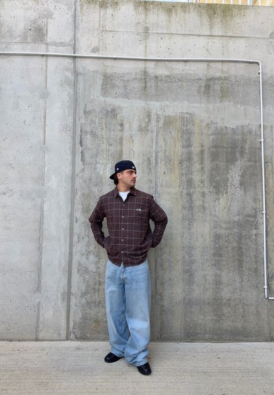 Camisa de cuadros marrón con botones, jeans sueltos azul claro y zapatos negros. Fondo de pared de hormigón con textura y líneas visibles.