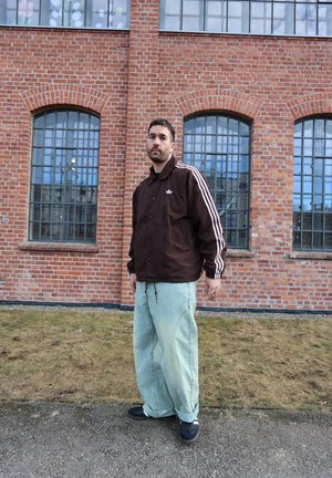 Hombre con chaqueta marrón de Adidas y jeans sueltos de color azul claro de pie frente a un edificio de ladrillos rojos con grandes ventanas arqueadas.