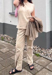 Sieviete, kas stāv uz ietves, valkājot bēšu krāsas bezroku kreklu un bikses, tur brūnu jaku un nēsā rozā somiņu, uz kājām melnas sandales.