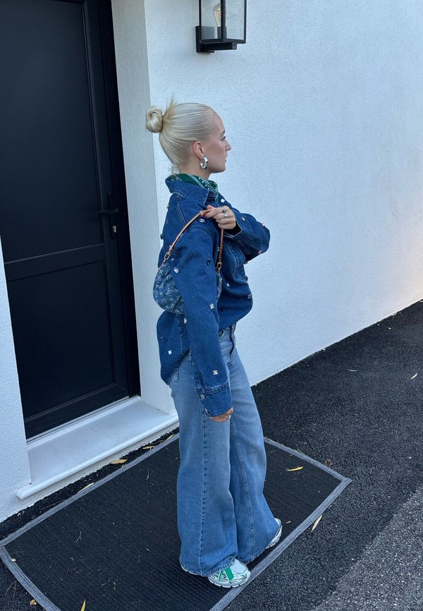 Denimjakke med udsmykkede detaljer, grøn skjorte, brede jeans og mønstrede sneakers. En lille taske hænger over skulderen.