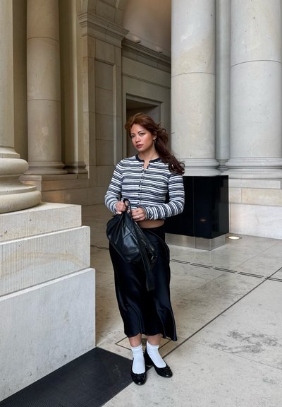 Joven mujer con cárdigan a rayas, falda negra, calcetines blancos y zapatos negros, sosteniendo una bolsa negra, de pie dentro de un gran edificio de piedra con columnas.