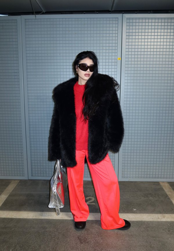 Frau trägt schwarzen Pelzmantel, roten Pullover, knallrote Hosen, schwarze Schuhe, große Sonnenbrille, hält eine silberne Handtasche und steht auf einem Parkplatz.