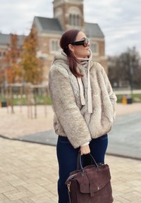 Jacke aus Kunstfell in Hellbeige mit Kapuze und Kordeln, kombiniert mit dunkelblauen Jeans und einer dunkelbraunen Wildledertasche.