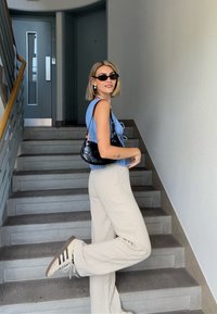 Top azzurro chiaro a costine, pantaloni a gamba larga beige, borsa nera, occhiali da sole neri, in piedi su scale grigie con sfondo di pareti chiare.