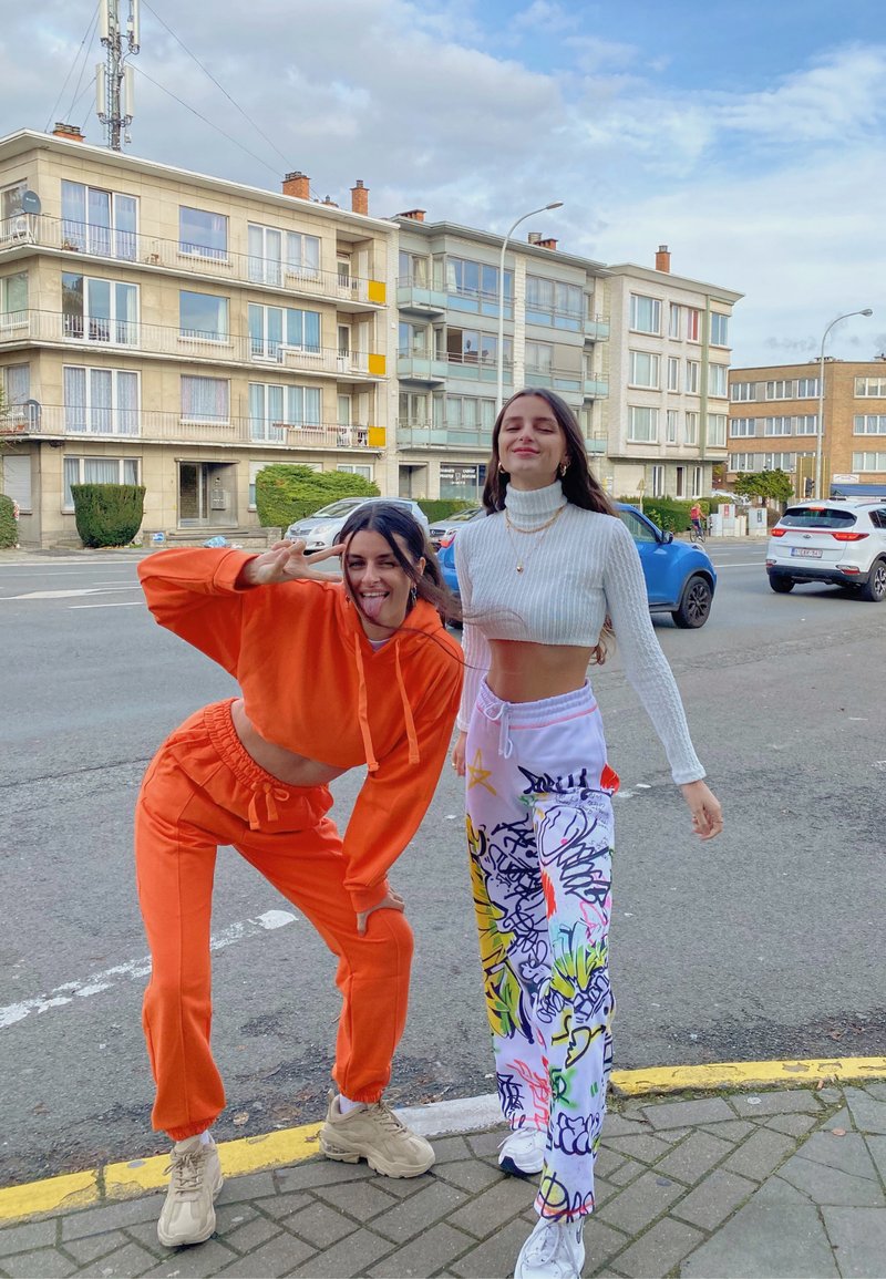 To personer poserer udendørs. Den ene har en orange cropped hoodie og matchende joggers på, mens den anden har en hvid ribbet turtleneck og mønstrede bukser.