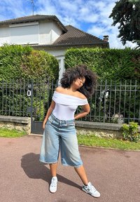 Body bianco scollato, culotte in denim blu chiaro, scarpe da ginnastica bianche con dettagli verdi, ambiente all'aperto con vegetazione sullo sfondo.