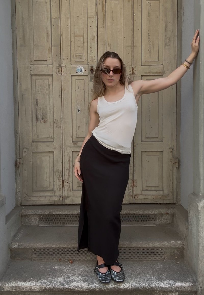 Junge Frau mit Sonnenbrille, die ein weisses Tanktop und einen langen schwarzen Rock trägt, posiert und lehnt an einer abgenutzten Holztür auf Steinstufen.