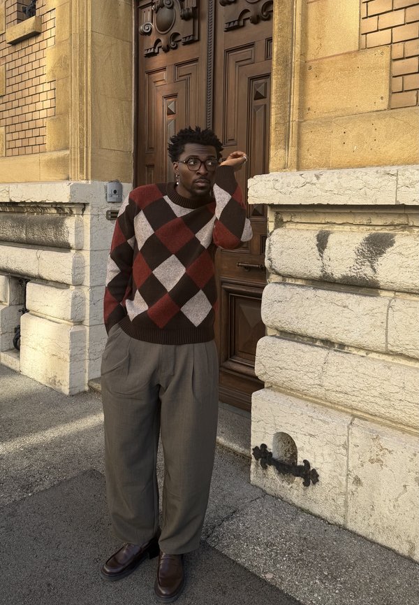 Homme portant des lunettes, un pull à losanges et un pantalon ample, appuyé contre un mur en pierre à côté d'une porte en bois dans une rue de la ville.