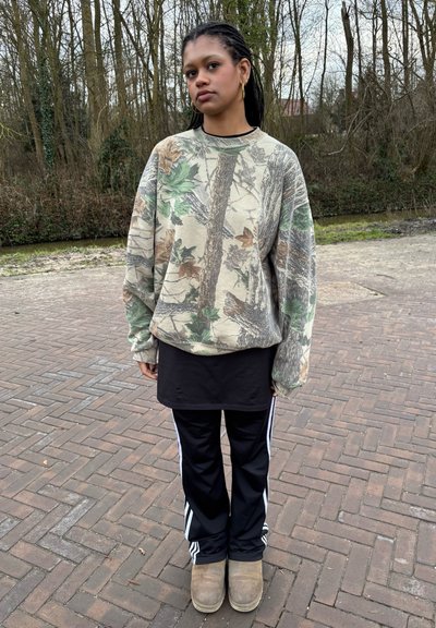 Sudadera oversized de camuflaje en beige con acentos en verde y marrón, combinada con una túnica negra y pantalones deportivos negros con rayas blancas.