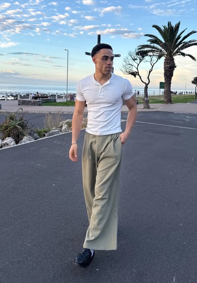 Joven con camisa blanca de manga corta y pantalones beige caminando por un sendero pavimentado cerca de palmeras, con el océano y un cielo nublado al fondo.