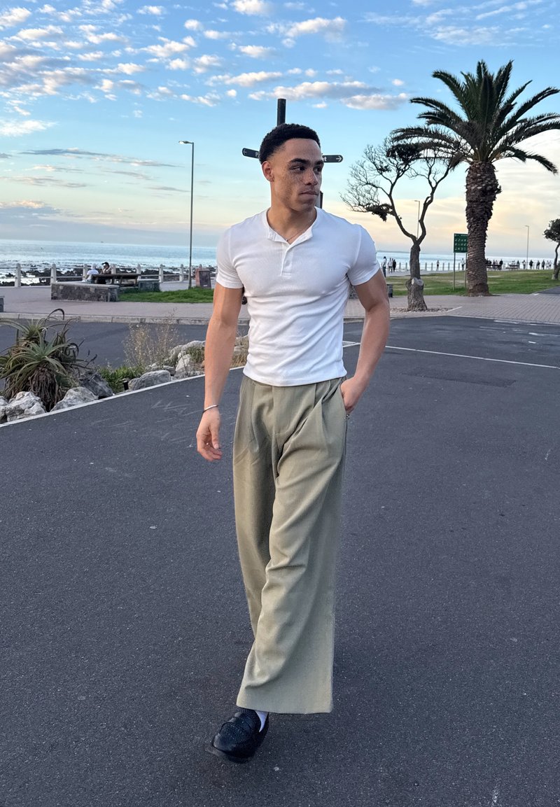 Νέος άνδρας με λευκό κοντομάνικο πουκάμισο και μπεζ παντελόνι περπατά σε στρωμένο μονοπάτι κοντά σε φοίνικες, με θέα στον ωκεανό και συννεφιασμένο ουρανό στο παρασκήνιο.