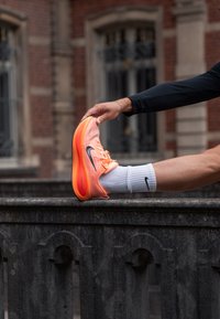 Buty biegowe Nike Zoom w pomarańczowo-różowym gradiencie, z siateczkową cholewką, czarnym logo Swoosh, noszone w białej skarpecie, stopa rozciągająca się na kamiennej półce.