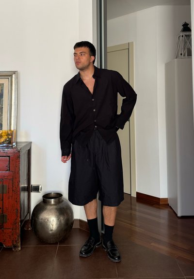 Camisa negra de botones con corte holgado, combinada con pantalones cortos negros con cordón. Medias y zapatos negros, de pie al lado de un jarrón metálico decorativo.