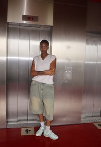 Top smanicato bianco a coste, pantaloni corto di denim azzurro, sneakers verde menta. In piedi davanti a un ascensore metallico con pavimento rosso.