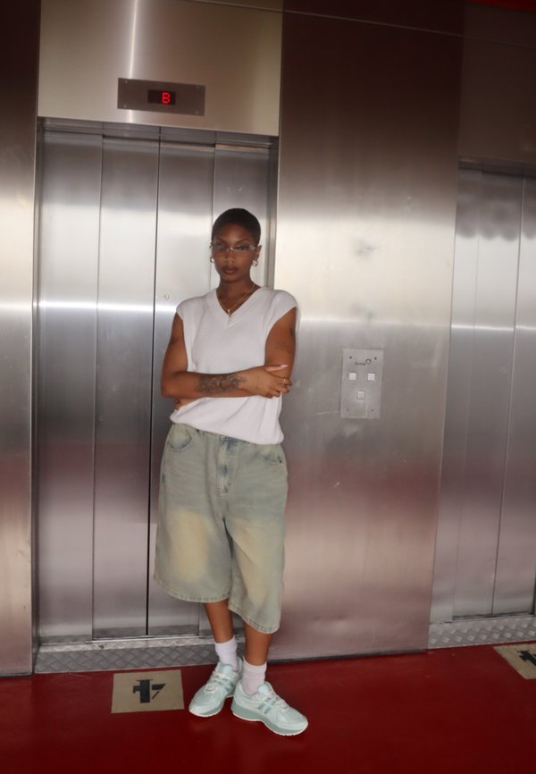 Top smanicato bianco a coste, pantaloni corto di denim azzurro, sneakers verde menta. In piedi davanti a un ascensore metallico con pavimento rosso.