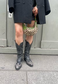 Botas de cowboy em couro preto com costuras brancas intrincadas e detalhes em estampado de cobra. Acompanhadas de uma pequena bolsa estampada em bege e verde.