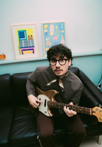 Camisa listada a castanho e um cachecol; segurando uma guitarra elétrica dourada com um escudo branco. Sofá de couro preto; obras de arte coloridas emolduradas ao fundo.