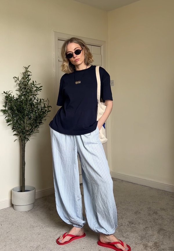 Vrouw draagt een zwart T-shirt, losse witte broek met fijne strepen, rode slippers, zonnebril en draagt een beige handtas binnen in de buurt van een kamerplant.