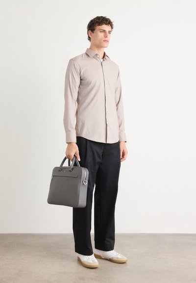 Maletín de cuero gris con asas superiores, combinado con una camisa de botones beige clara y pantalones azul marino. El modelo lleva zapatillas blancas.