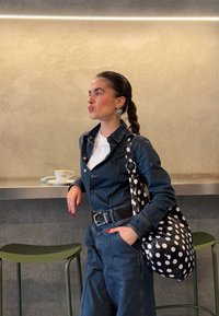 Γυναίκα με πλεξούδες που φοράει σκούρο τζιν μπουφάν και παντελόνι, στέκεται δίπλα σε έναν πάγκο με ένα φλιτζάνι καφέ, κρατώντας μια μαύρη τσάντα με πουά σχέδιο.