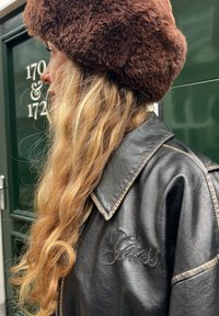 Femme aux longs cheveux blonds portant un chapeau marron duveteux et une veste en cuir noire de la marque Guess, debout devant des portes vertes avec des numéros de maison.