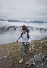 Person, die ein gestreiftes weißes und graues Hemd, olivgrüne Hose und farbenfrohe Wanderschuhe trägt, während sie einen schwarzen Rucksack in einer felsigen Landschaft trägt.