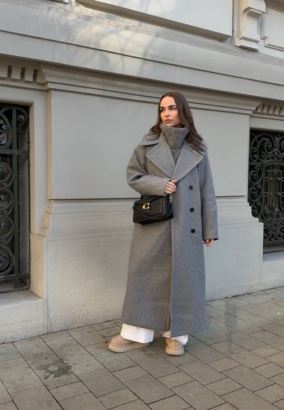 Mujer con un abrigo largo gris, suéter de cuello alto gris, pantalones blancos, zapatos beige y un bolso cruzado negro, de pie sobre una acera pavimentada.