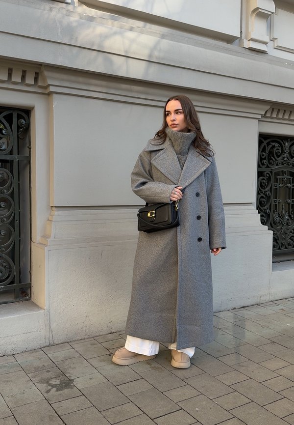 Femme portant un long manteau gris, un pull à col roulé gris, un pantalon blanc, des chaussures beige et un sac noir en bandoulière, debout sur un trottoir pavé.