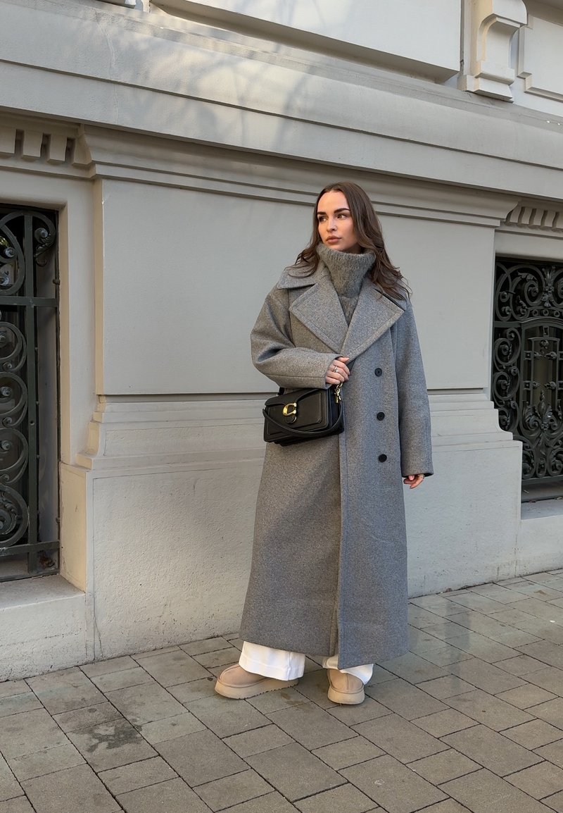 Femme portant un long manteau gris, un pull à col roulé gris, un pantalon blanc, des chaussures beige et un sac noir en bandoulière, debout sur un trottoir pavé.