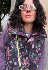 Γυναίκα με σγουρά σκούρα μαλλιά που φοράει ανοιχτό μπεζ γυαλιά ηλίου, μοβ γιλέκο με λουλουδάτο σχέδιο και λιλά πουλόβερ, στέκεται μπροστά σε μια μπεζ τοίχο.