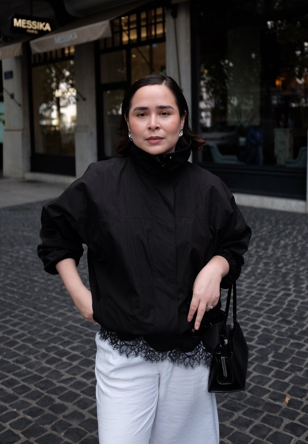 Mustavärinen, kevyt bomber-takki korkealla kauluksella, joka on puettu valkoisen pitsireunuksilla varustetun asun päälle. Pieni musta käsilaukku ja helmikorvakorut näkyvissä.