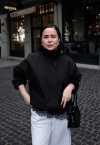 Mustavärinen, kevyt bomber-takki korkealla kauluksella, joka on puettu valkoisen pitsireunuksilla varustetun asun päälle. Pieni musta käsilaukku ja helmikorvakorut näkyvissä.