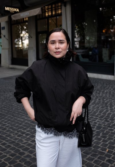 Chaqueta bomber negra y ligera con un cuello alto, llevada sobre un conjunto blanco con puntilla. Se puede ver un pequeño bolso negro y pendientes de perlas.