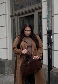 Clutch castanha em camurça com um formato estruturado e alças finas, apresentando um pequeno detalhe em prateado minimalista; usada por uma pessoa com um trench coat castanho.