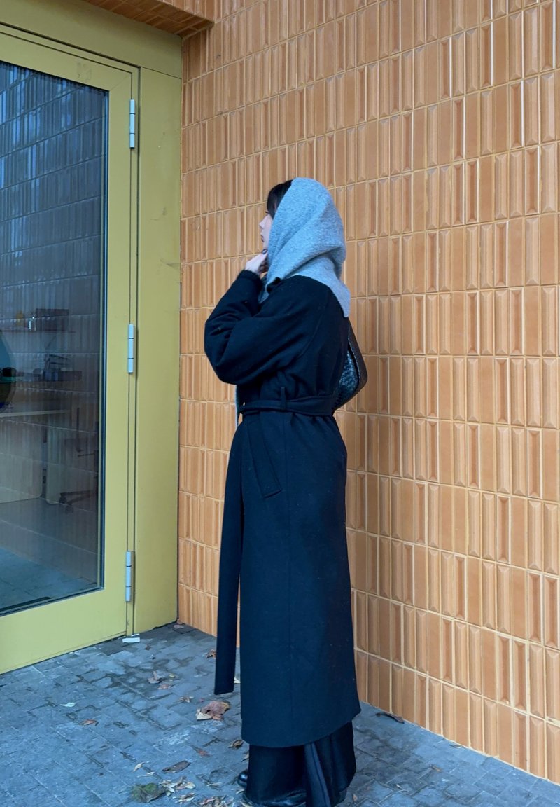 Manteau long noir avec une ceinture nouée à la taille, porté par-dessus une longue robe noire. Capuche grise. Debout à côté d'une porte en verre contre un mur texturé en carrelage orange.