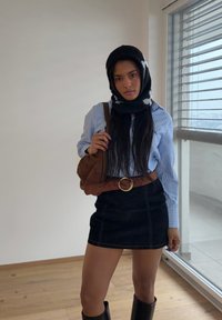 Jeune femme portant un foulard noir à motifs, chemise rayée bleue, jupe noire, ceinture marron, sac marron, debout à l'intérieur près d'une fenêtre.