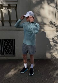 Lichtgroene lange mouw top, grijze shorts, witte pet, zwarte hardloopschoenen met blauwe accenten, terwijl je de pet aanpast, staand in het zonlicht.