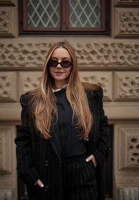 Γυναίκα με μακριά ξανθά μαλλιά που φοράει μαύρα γυαλιά ηλίου σχήματος γάτας, μαύρο παλτό με ρίγες και ταιριαστό παντελόνι στέκεται μπροστά σε έναν τοίχο με υφή από πέτρα.