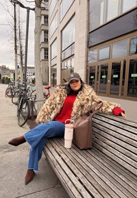 Manteau en fausse fourrure, pull rouge et jean évasé bleu. Sac à main marron à côté d'une tasse de voyage rose avec anse, posés sur un banc en bois.