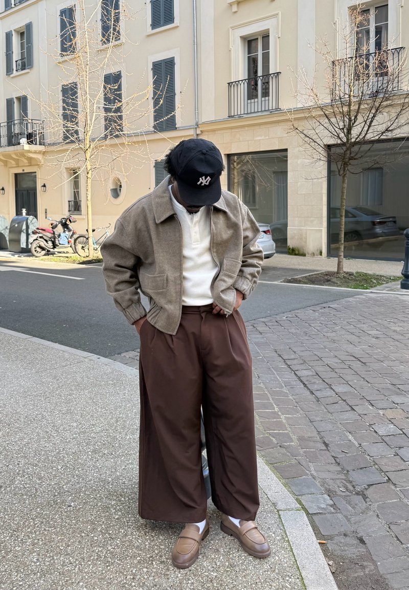 Άτομο που φοράει μαύρο καπέλο, μπεζ σακάκι, άσπρο πουκάμισο, φαρδιά καφέ παντελόνια και καφέ παπούτσια, που στέκεται σε πεζοδρόμιο σε αστική οδό.