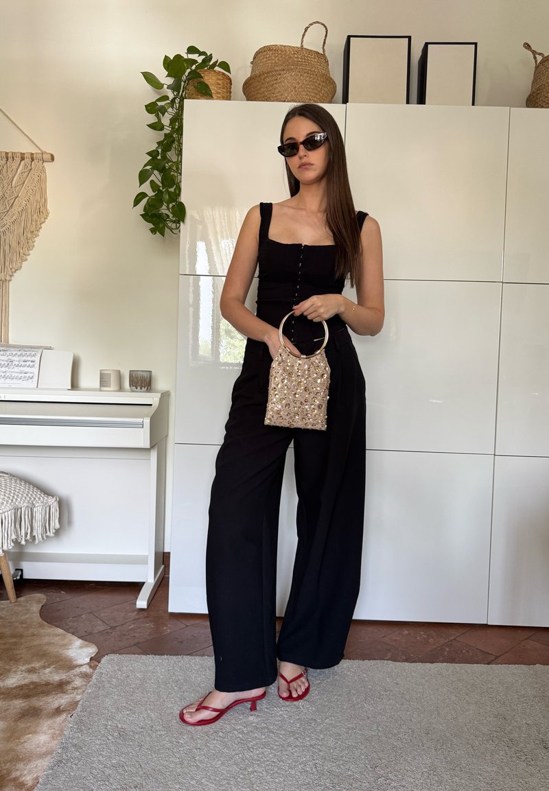 Femme en combinaison noire et talons rouges tenant un sac à main doré décoratif, portant des lunettes de soleil, debout à l'intérieur près d'armoires blanches et plantes.
