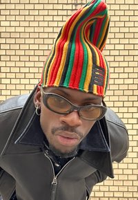 Virkat mössa med hög, spetssig topp. Har växelvis röda, gröna och gula ränder med en svart accent. Glansiga svarta solglasögon.