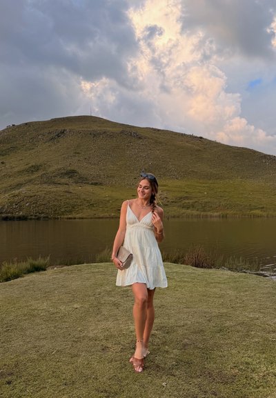 Joven mujer con vestido blanco sosteniendo un bolso de mano, de pie en la orilla con hierba junto a un lago con colinas onduladas y cielo nublado de fondo.